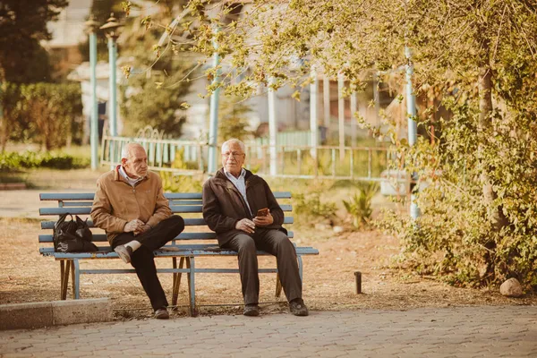 Two senior men sitting and conversing on a bench in a sunny autumn park.