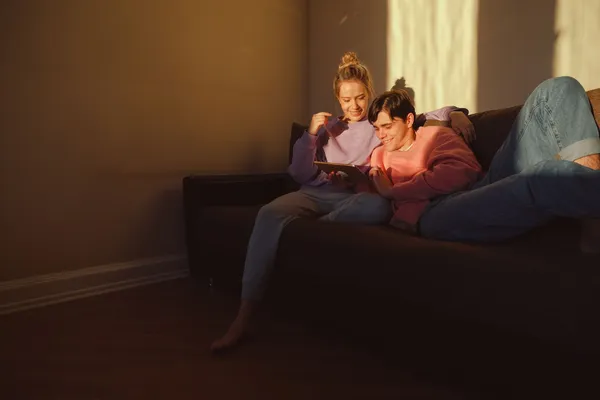 A couple sits on a sofa using a digital tablet in warm evening light, enjoying a quiet moment together.