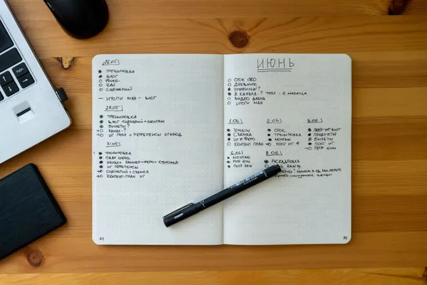 An open notebook with handwritten notes resting on a wooden desk beside a laptop and pen.