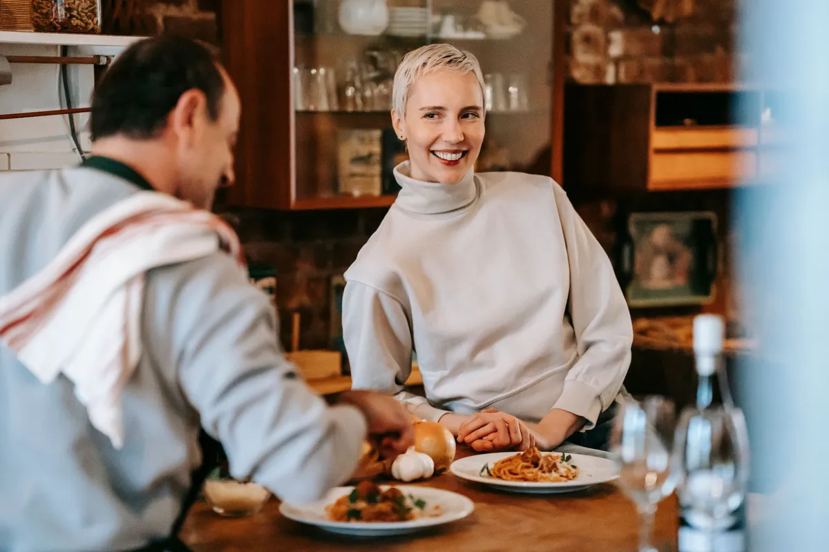 Smiling adult couple in casual clothes having dinner while male cooking pasta on plates at counter near glasses and wine bottle near various products and ingred