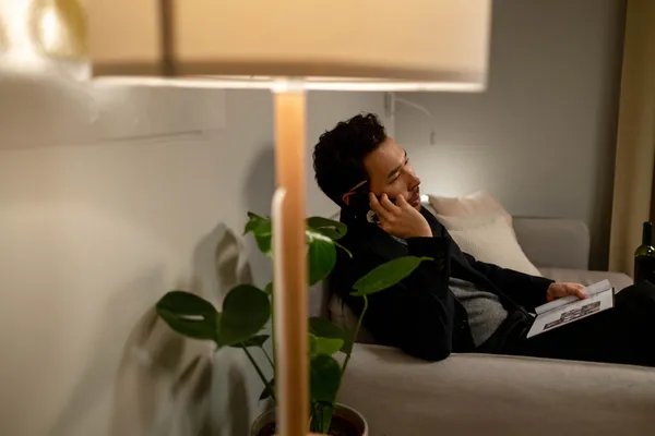 A man relaxing on a sofa indoors, talking on a cellphone with a book. Perfect cozy home ambiance.
